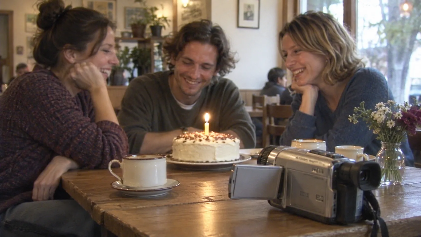 Concept illustration of three adult friends around a birthday cake with a compact silver camcorder in the foreground